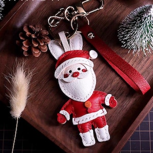 Könnte beinhalten: Ein Schlüsselanhänger aus Leder in Form einer Weihnachtsmannfigur mit Hasenohren, mit einer rot-weißen Farbgebung, genähten Details, einem goldfarbenen Metallring und einer passenden roten Lederhandschlaufe an der Oberseite der Figur.