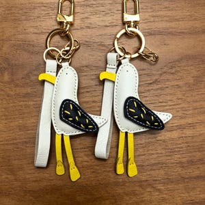 May include: Two white leather seagull keychains with gold-colored hardware. The seagulls have black and yellow wing details and yellow legs. Each keychain has a white leather strap and a gold-colored clasp.
