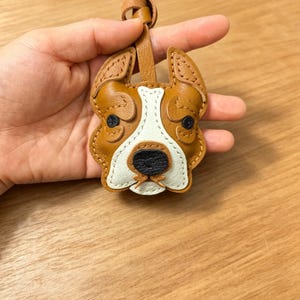 May include: A tan and white leather dog head keychain featuring stitched details, dark brown eyes, and a black nose. The dog-shaped charm has a leather loop at the top for attachment, showcasing layered leather construction with visible stitching.