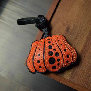 May include: A pumpkin-shaped leather charm featuring a textured orange surface with black polka dots. The segments are defined by visible yellow stitching, and it includes a black leather loop strap for attachment to bags or other personal items.