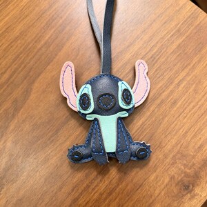 May include: A dark blue leather-like keychain shaped like the character Stitch, featuring light blue accents on the chest and around the eyes, pale pink inner ears, and dark blue stitching throughout. A long, thin dark blue strap is attached to the top.