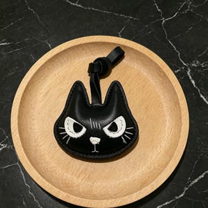 Può includere: Un portachiavi nero a forma di gatto con dettagli ricamati bianchi, tra cui occhi, baffi e naso. Il portachiavi ha un cinturino in pelle nera ed è appoggiato su un piatto di legno.