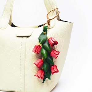Op de afbeelding: Een crèmekleurige handtas met textuur en een decoratieve leren hanger in de vorm van een tros rode tulpbloesems en groene bladeren. De hanger is aan het handvat bevestigd met een dun beige leren koordje en een kleine messingkleurige klinknagel.