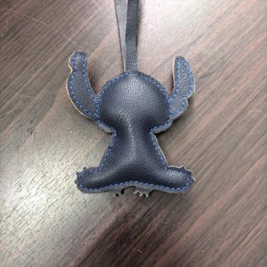 May include: A dark navy blue faux leather keychain shaped like a cartoon character with large ears and a rounded head. The item features visible blue stitching along the edges and includes a matching dark navy blue leather strap for hanging.
