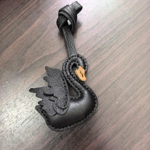 May include: A black textured leather swan charm featuring a knotted strap at the top. The swan has a distinct layered wing design and a small, light tan leather patch on its beak. The edges are finished with visible black stitching throughout.