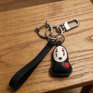 May include: A black leather keychain with a silver clasp and key rings. The keychain features a black and white cartoon character with a red heart. The keychain is on a wooden surface.