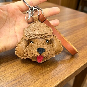 May include: A brown leather poodle-shaped keyring. The dog's face has a black nose and a red tongue. The keyring includes a brown leather strap and a silver metal ring.