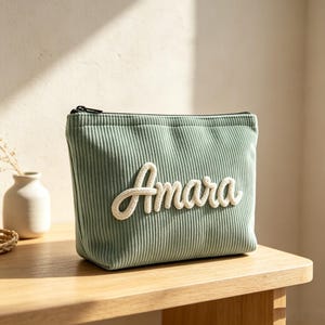 May include: A sage green corduroy zippered pouch featuring the name Amara in white, raised, cursive rope-style lettering centered on the front. The fabric has a distinct vertical ribbed texture, and the pouch has a black zipper closure at the top.