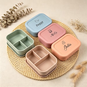 Puede incluir: Una colección de fiambreras rectangulares de silicona en tonos pastel azul, rosa y melocotón, con tapas personalizadas con los nombres Peggy, Molly y Julia junto a pequeños iconos. También se ven dos recipientes abiertos en verde salvia y rosa.