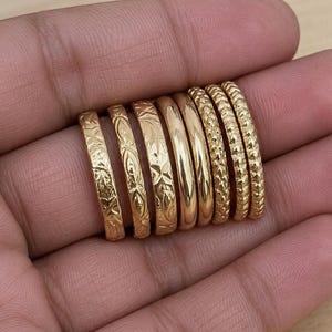 Könnte beinhalten: Eine Sammlung von acht goldfarbenen Ringen, die in einer Reihe angeordnet sind. Die Ringe weisen verschiedene Designs auf, darunter drei mit komplexen Gravurmustern, zwei mit glatten, polierten Oberflächen und drei mit einem texturierten Muster.