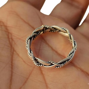 Könnte beinhalten: Ein silberner Ring mit gedrehtem Design, der ein glattes, poliertes Band mit einem dünneren, seilartig strukturierten Strang kombiniert. Das Metall hat eine helle, reflektierende Oberfläche mit abgedunkelten Vertiefungen, die das Flechtmuster betonen.