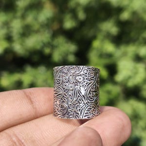 Könnte beinhalten: Ein breiter silberfarbener Ring mit einem detaillierten, geprägten Blumen- und Rankenmuster auf der gesamten Oberfläche. Das Metall weist in den vertieften Bereichen ein dunkles, oxidiertes Finish auf, das die Gravuren hervorhebt.