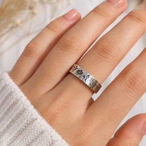 Könnte beinhalten: Ein silberfarbener Metallring mit einer Reihe kleiner, schwarz gravierter Blumen- und Pflanzenmotive auf der Außenseite. Der Ring hat ein flaches Profil und eine glatte, polierte Oberfläche, die den Finger mit gleichmäßiger Breite umschließt.