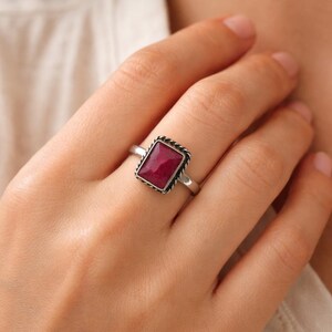 Könnte beinhalten: Ein Silberring mit einem rechteckigen, tief rosa-roten Edelstein, der in eine gedrehte, seilartige Metallfassung eingefasst ist. Die Ringschiene ist glatt und poliert und hält den Stein sicher in einer Zargenfassung, die die Farbe betont.