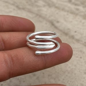 Könnte beinhalten: Ein silberfarbener Bypass-Ring mit einem mehrschichtigen Spiraldesign. Das Band besteht aus drei einzelnen, überlappenden Windungen, die sich um den Finger legen und jeweils in einer glatten, abgerundeten Spitze mit polierter, reflektierender Oberfläche enden.
