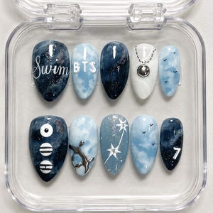 May include: A set of ten oval press-on nails in a clear case. Designs include the words "Swim" and "BTS", celestial patterns, and the number 7. The nails are navy, white, and light blue.