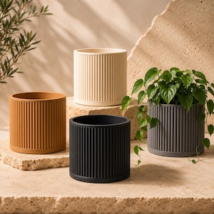 Op de afbeelding: Een set van vier cilindrische plantenpotten met een verticaal geribbelde textuur. De potten zijn afgewerkt in effen matte kleuren, waaronder crème, mosterdgeel, antracietgrijs en zwart. Eén grijze pot bevat een groene hangplant.