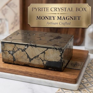 Könnte beinhalten: Eine rechteckige Schatulle mit einer metallischen, mosaikartigen Oberfläche und dunklen, unregelmäßigen Adern. Eine goldfarbene Plakette darüber zeigt den Text: PYRITE CRYSTAL BOX, MONEY MAGNET, Artisan Crafted. Die Schatulle steht auf einem Holzbrett.