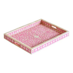 Könnte beinhalten: Ein rechteckiges Serviertablett mit einer leuchtend rosa Basis und einem komplizierten weißen Blumenrankenmuster. Das Tablett hat erhöhte Seiten mit integrierten Griffen und ein sich wiederholendes weißes geometrisches Rautenmuster am Rand.