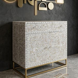 May include: A rectangular cabinet featuring an intricate floral mother-of-pearl inlay pattern on a light grey surface. The piece includes one top drawer and two lower doors with clear knobs, supported by a gold-toned metal frame base.