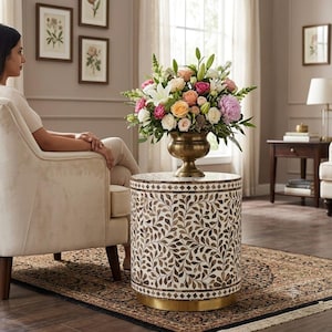 Könnte beinhalten: Ein zylindrischer Beistelltisch mit einem komplizierten eingelegten Blattmuster in Braun- und Cremetönen, gekrönt von einer messingfarbenen Standvase, die mit einem lebendigen Blumenarrangement aus rosa, pfirsichfarbenen, weißen und grünen Blüten gefüllt ist.