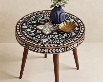 Op de afbeelding: Een ronde bijzettafel met drie taps toelopende houten poten heeft een zwart blad ingelegd met witte bloemmotieven en een centraal bloemmotief van parelmoer. Een blauwe geribbelde vaas met een groene plant en een klein gouden schaaltje staan erop.