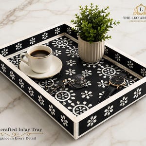 Könnte beinhalten: Ein rechteckiges schwarzes Tablett mit einem weißen Blumen-Intarsienmuster auf der Basis und den Seiten. Das Tablett enthält eine weiße Kaffeetasse mit Untertasse, eine kleine grüne Pflanze in einem gerippten Topf, eine Brille und eine schwarze Armbanduhr.