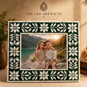 May include: A square picture frame featuring a dark green border with an inlaid white floral pattern made of mother-of-pearl. The frame displays a photograph of two children sitting by a lake. The text THE LEO ARTIFACTS HANDMADE WITH ELEGANCE is visible.