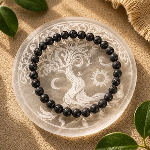 Peut inclure: Un bracelet de perles en pierre de lave noire repose sur une plaque décorative ronde blanche. La plaque présente un motif d'arbre de vie avec un croissant de lune et un soleil. Le bracelet est fait de petites perles sombres et rondes.