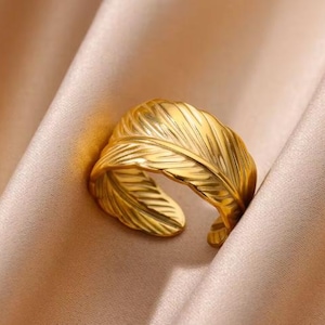 May include: A gold-colored adjustable ring with a feather design. The ring features detailed feather vein textures and is set against a soft, neutral-toned background. The ring is made of a shiny, reflective metal.