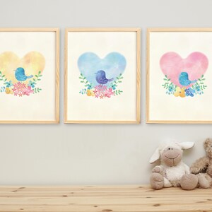 以下が含まれることがあります： 黄色、青、ピンクのハートの中に青い鳥が描かれた水彩画風の壁掛けアートプリント3枚セット。それぞれのハートの基部にはカラフルな花の装飾が施されており、明るい木製のフレームに収められて壁に飾られています。
