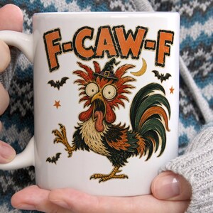 May include: A white ceramic mug featuring a colorful graphic of a wide-eyed rooster wearing a small witch hat. The rooster has red, black, and orange feathers. Above the rooster, the text F-CAW-F is printed in orange. Small black bats and orange stars surround the central rooster illustration.