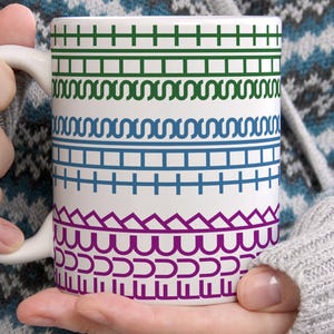May include: A white ceramic mug featuring horizontal bands of geometric patterns in green, blue, and purple. The design includes rows of crosses, squares, and scalloped lines that wrap around the exterior surface of the cylindrical drinking vessel.