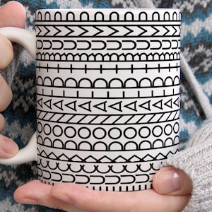 May include: A white ceramic mug featuring a repeating black graphic pattern of horizontal bands. The design consists of various geometric shapes including circles, triangles, semicircles, and lines arranged in distinct rows around the exterior surface.