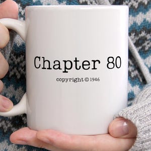 May include: A white ceramic mug featuring the black printed text Chapter 80 and the smaller text copyright 1946 centered on the front. The mug has a smooth cylindrical body and a curved handle on the side, designed for holding hot or cold beverages.