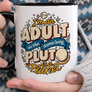 May include: A white ceramic mug with a black handle and rim featuring a colorful graphic print that reads I'm an ADULT in the same way PLUTO is a Planet, surrounded by illustrations of stars, a comet, and a moon against a white background.