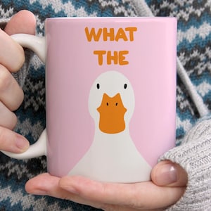 May include: A light pink ceramic mug featuring a graphic of a white duck with an orange beak. Above the duck, the text WHAT THE is printed in orange capital letters. The mug has a white handle and a smooth, cylindrical body design.