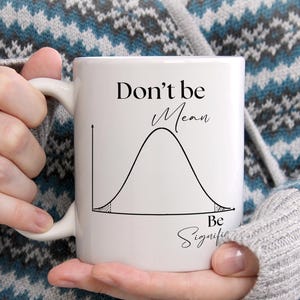 May include: A white ceramic mug featuring a black line drawing of a bell curve distribution graph. The text "Don't be Mean" is printed above the curve, and the text "Be Significant" is printed below the curve in a black script font.