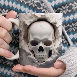 May include: A white ceramic mug featuring a three-dimensional relief design of a human skull emerging from a cracked surface surrounded by sculpted floral elements. The skull is centered on the mug, with a smooth handle on the left side.