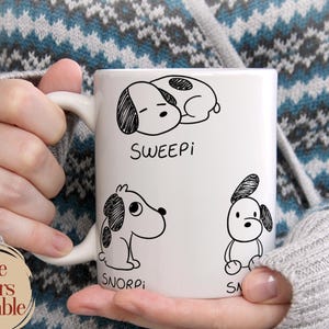 May include: A white ceramic mug featuring three black line-art illustrations of cartoon dogs. The top dog is labelled SWEEPi, the bottom left dog is labelled SNORPi, and the bottom right dog is labelled SA. The mug has a handle on the left side.