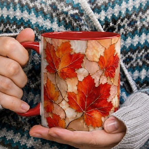 May include: A ceramic mug with a glossy red interior and handle, featuring a vibrant exterior pattern of overlapping autumn maple leaves in shades of orange, red, and cream, outlined with thin gold lines that create a stained glass effect.