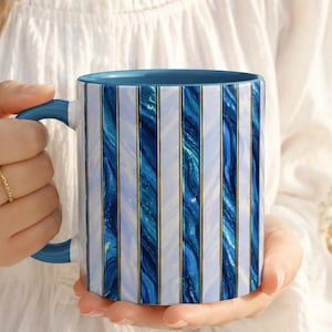 May include: A ceramic mug featuring a vertical striped pattern in shades of dark blue, light blue, and white with thin gold lines. The mug has a solid blue handle and a matching blue interior. A circular badge on the left reads More Colors Available.