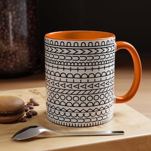 May include: A ceramic mug with a bright orange interior and handle, featuring a white exterior decorated with horizontal bands of black geometric patterns including circles, triangles, lines, and semicircles arranged in a repeating sequence.