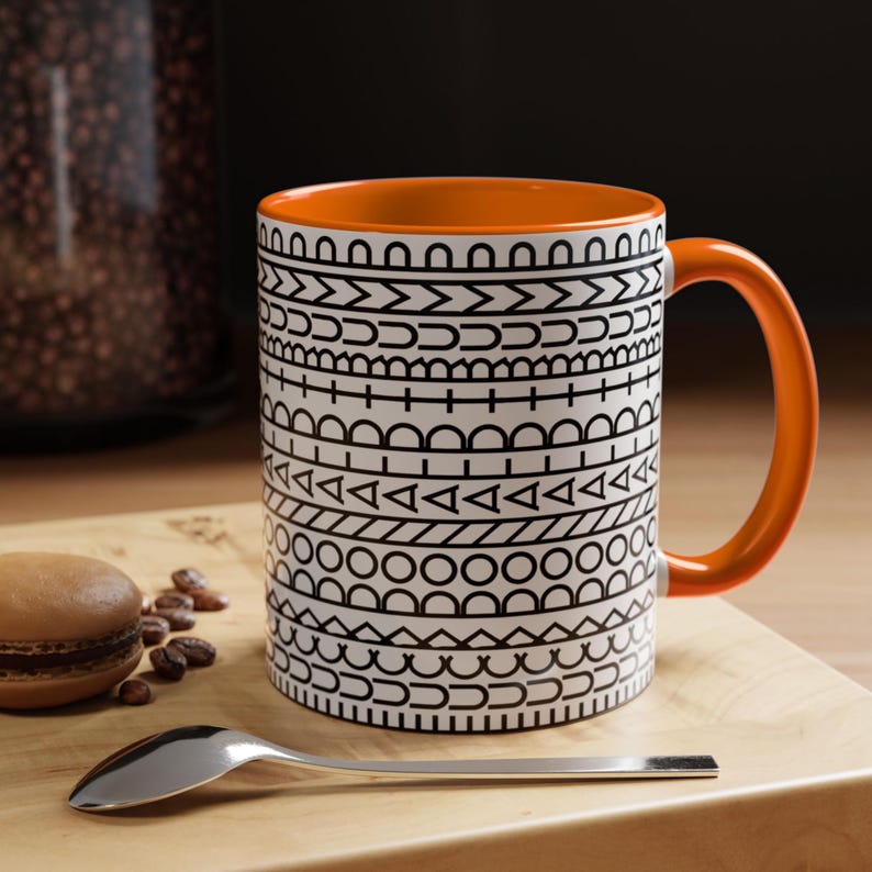 May include: A ceramic mug with a bright orange interior and handle, featuring a white exterior decorated with horizontal bands of black geometric patterns including circles, triangles, lines, and semicircles arranged in a repeating sequence.