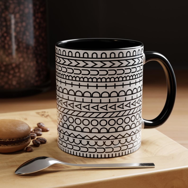 May include: A white ceramic mug with a black interior and handle, featuring a repeating horizontal pattern of black geometric shapes including circles, triangles, lines, and arches stacked in distinct rows around the exterior surface.