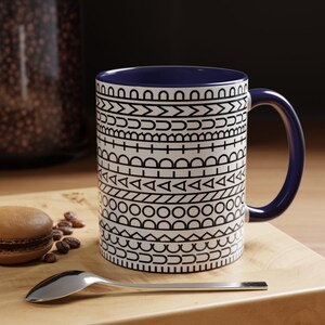 May include: A ceramic mug with a dark blue interior and handle, featuring a white exterior decorated with horizontal bands of black geometric patterns including circles, triangles, lines, and semicircles arranged in a repeating sequence.