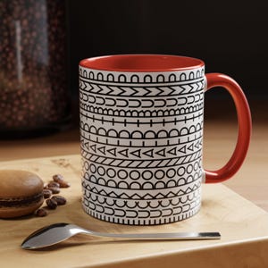 May include: A ceramic mug with a white exterior featuring horizontal bands of black geometric patterns, including circles, triangles, and lines. The interior of the mug and the curved handle are a solid, vibrant red colour, providing a contrast.