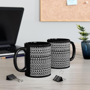 May include: Two black ceramic mugs featuring a repeating white geometric pattern of horizontal bands. The designs include lines, circles, triangles, and arches arranged in a stacked sequence around the exterior of each cylindrical mug.