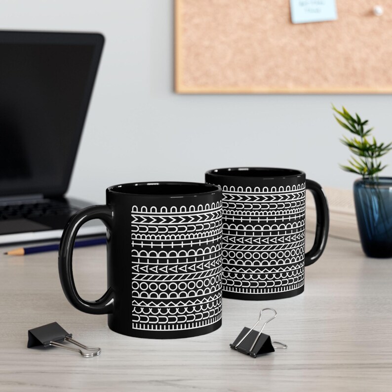 May include: Two black ceramic mugs featuring a repeating white geometric pattern of horizontal bands. The designs include lines, circles, triangles, and arches arranged in a stacked sequence around the exterior of each cylindrical mug.