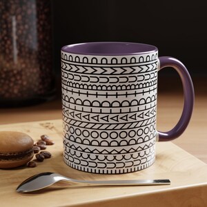 May include: A ceramic mug with a purple interior and handle, featuring a white exterior decorated with horizontal bands of black geometric patterns including circles, triangles, lines, and arches, arranged in a repeating, stacked design.
